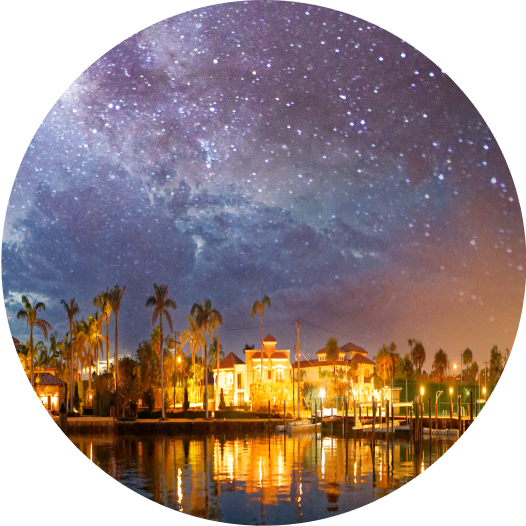 Nightime exterior of Boca Raton from the water. The sky shows stars, and the water in front reflects the lighted exterior at the port.